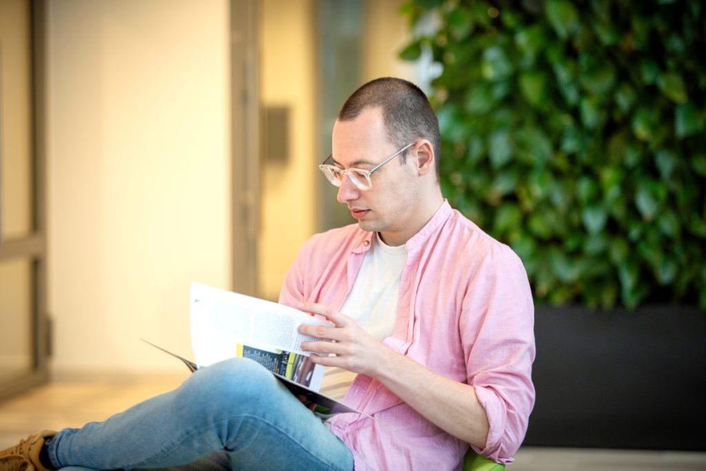 Een software engineer met een roze overhemd en bril zit op een groene stoel in de centrale hal bij Technolution, portret 2 van 5