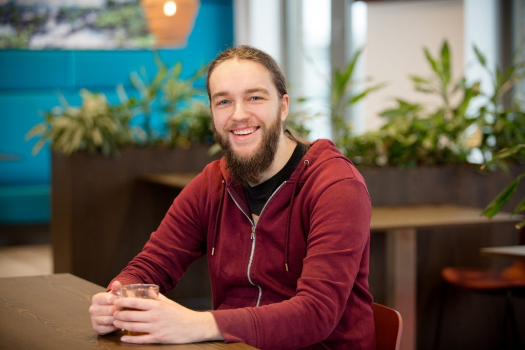 Een electrical engineer met een bordeauxrode hoodie zit ontspannen aan een tafel in kantoor bij Technolution portret 2 van 5