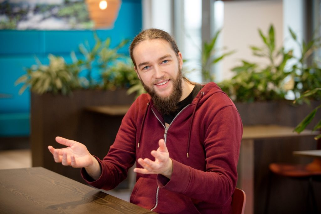 Een electrical engineer met een bordeauxrode hoodie zit ontspannen aan een tafel in kantoor bij Technolution portret 3 van 5