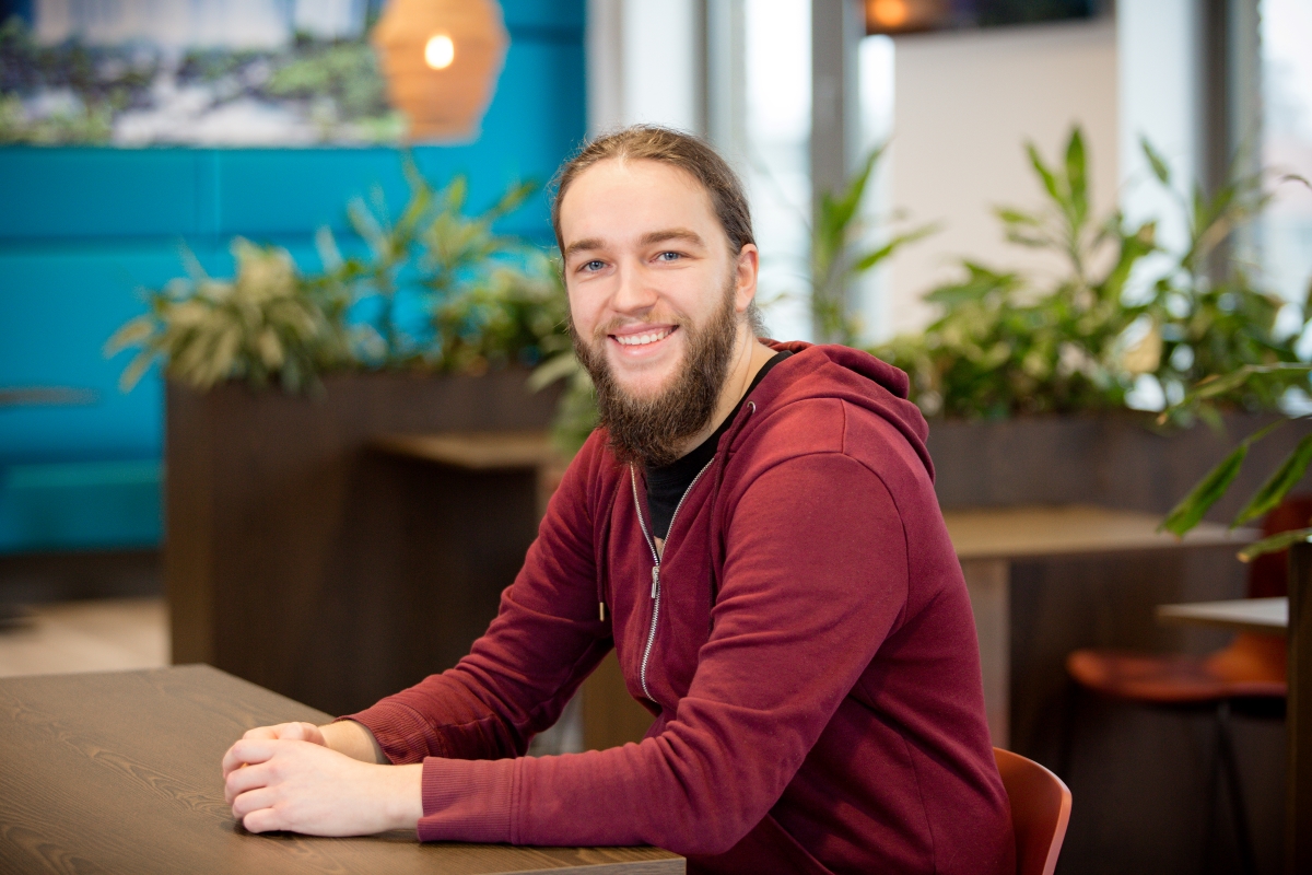 Een electrical engineer met een bordeauxrode hoodie zit ontspannen aan een tafel in kantoor bij Technolution portret 5 van 5